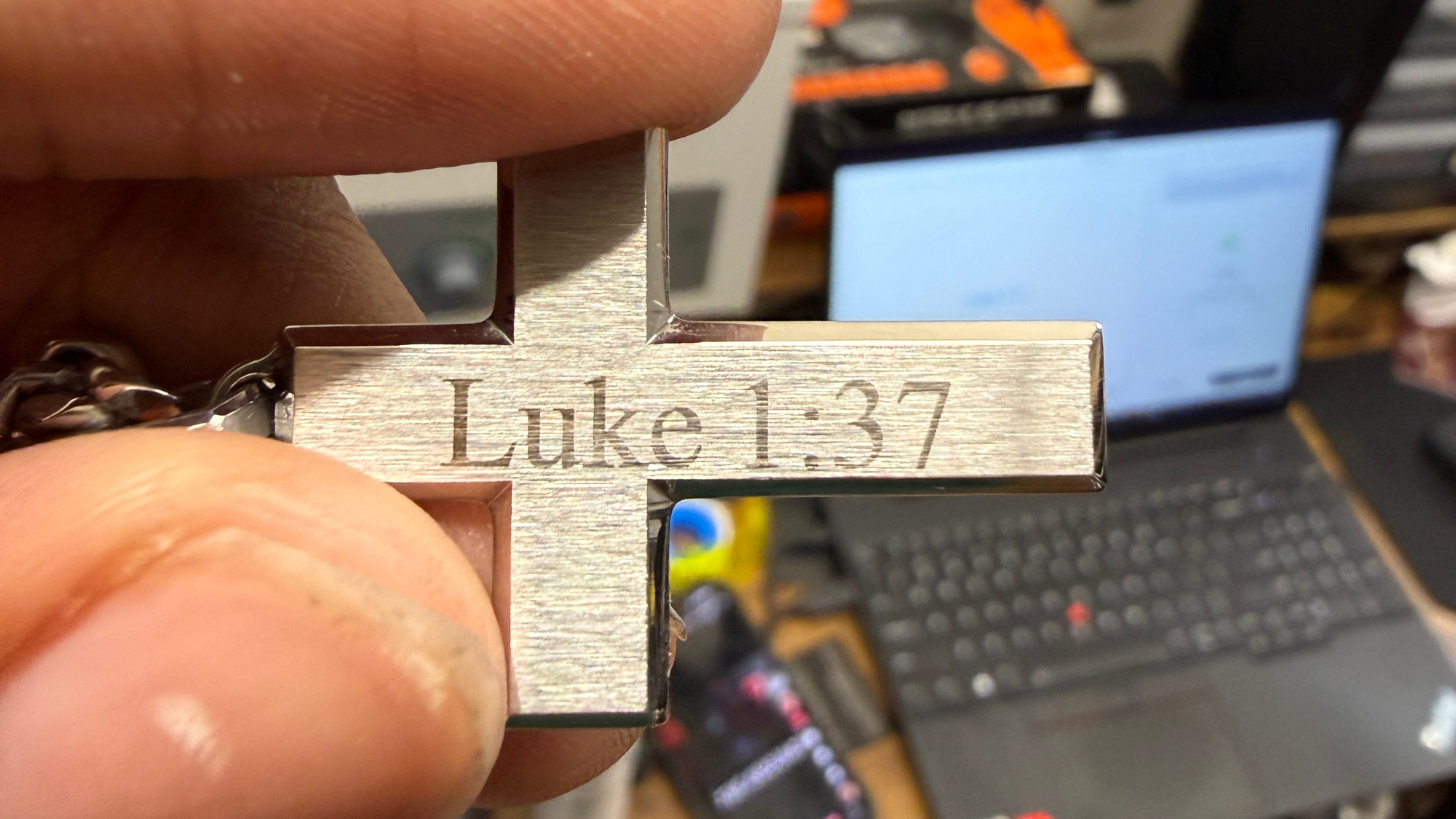 Metal cross with 'Luke 1:37' engraved, held in a hand with a blurred office background.